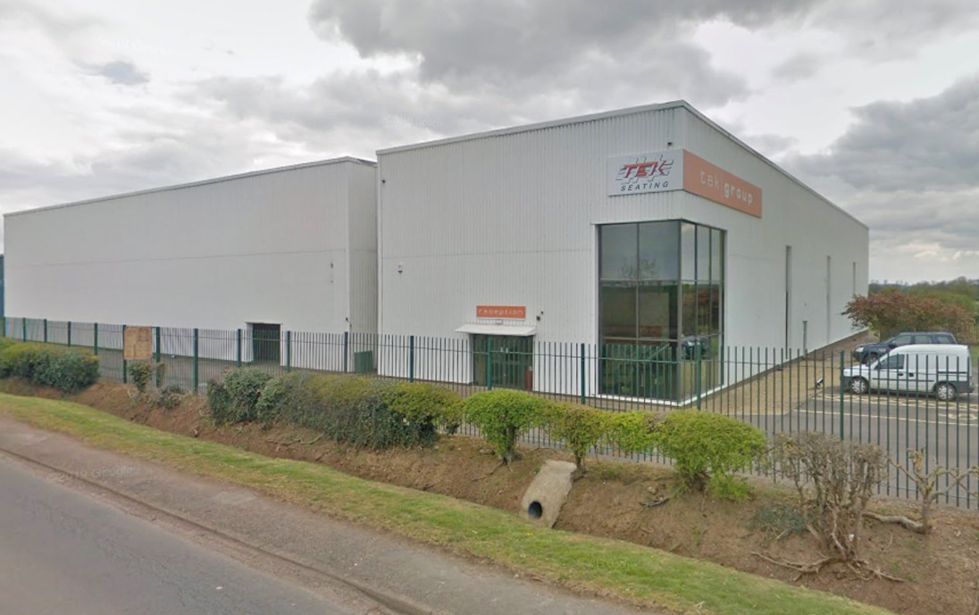 Exterior view of a modern white commercial warehouse building behind a metal security fence and roadside landscaping